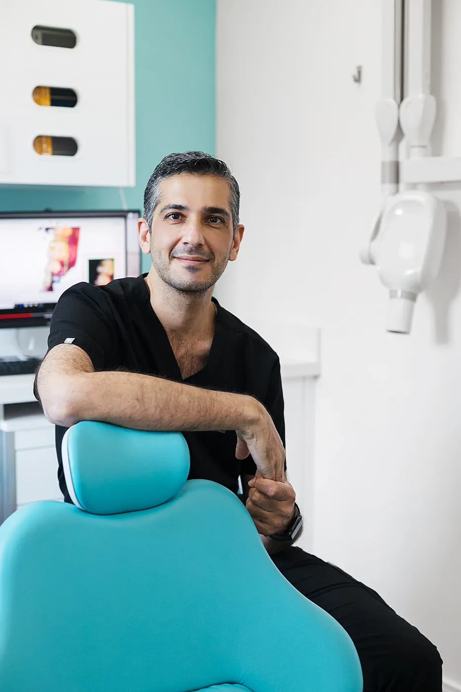 Dr. Mudhar seated beside the teal dental chair in the treatment room